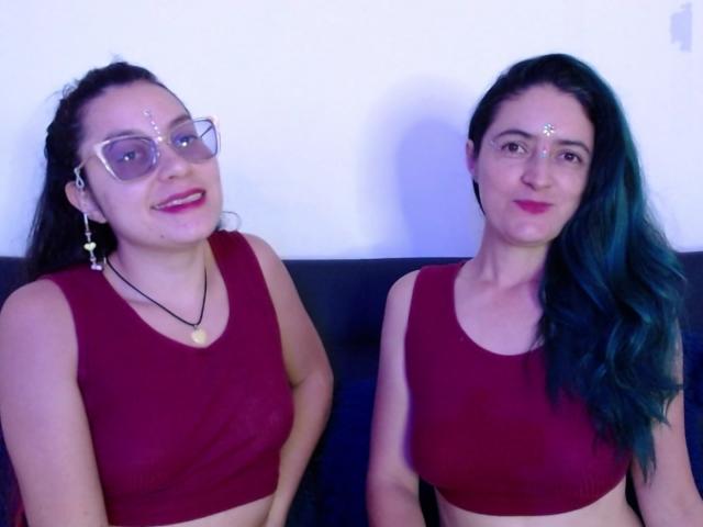 LunaVenus and LunaVenus, both with red crop tops, glasses, and forehead dots, sitting on a black couch. LunaVenus on left has dark hair, LunaVenus on right has green hair.