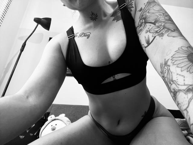 SujannaVarma with tattoos, wearing a black sports bra with a cutout and matching panties, showing her tits and a small tattoo above her left breast. Black-and-white selfie.
