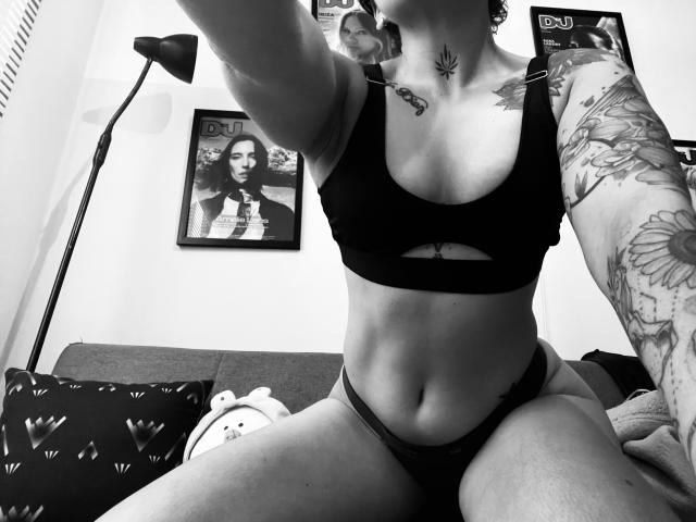 Black-and-white selfie of SujannaVarma with tattoos, wearing a sports bra and panties, showing her tits and pussy, arm tattoos, and a lamp in the background.
