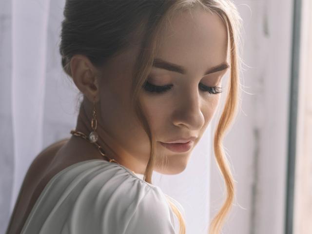 EvaDaly with blonde hair in a loose bun, wearing a white top, looking down with closed eyes, soft makeup, gold earrings, and a delicate necklace, in natural light.
