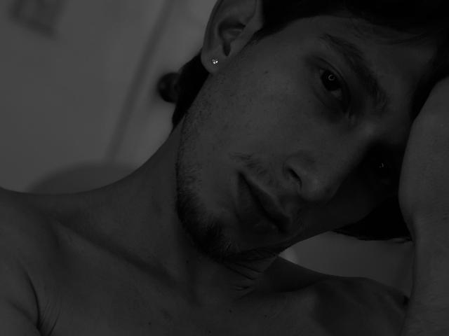DeimHt, shirtless, with a slight stubble, ear piercing, and a somber expression, rests his head on his arm in a black-and-white shot. His eyes are slightly downcast.