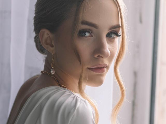 Eva Daly, com cabelos loiros ondulados, olhos azuis e maquiagem discreta, vestindo uma blusa branca, olhando por cima do ombro, usando brincos de ouro e um colar, em luz natural suave..