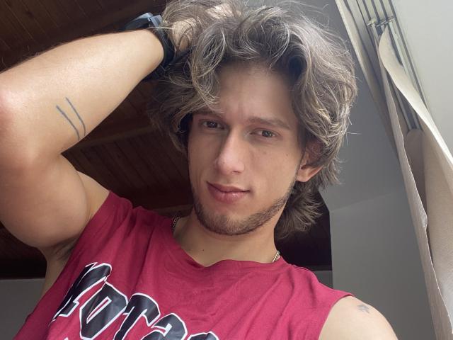 DeimHt with messy brown hair, a red sleeveless shirt, and a black wristband, raising his right arm, showing a tattoo and a slight beard, smiling at the camera.