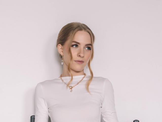 EvaDaly with blonde hair in a loose updo, wearing a white sheer top, small gold necklace, and earrings, looking to the side with a slight smile.