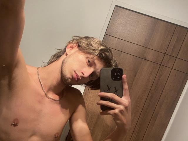 DeimHt with shoulder-length blonde hair, shirtless, wearing a necklace, holding a phone with a black case, nipple piercings, and a beard, taking a mirror selfie.
