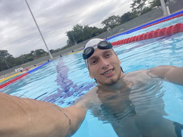 DeimHt, a muscular guy with a white swim cap and goggles, smiling in a pool, has a tattoo on his right arm and a visible penis underwater.