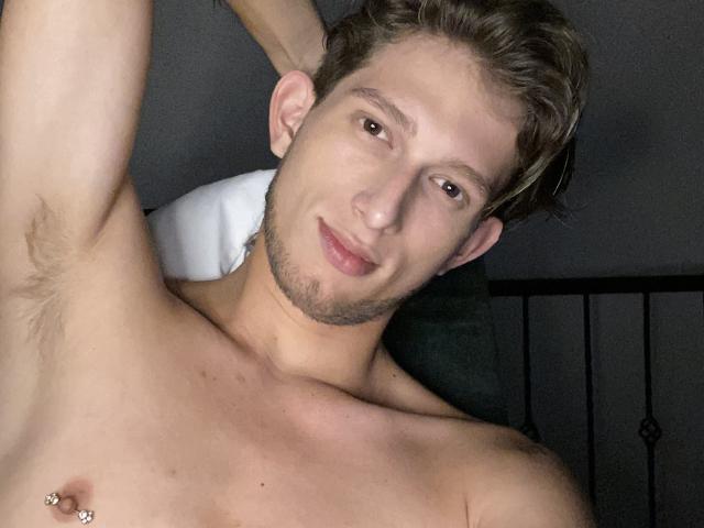 DeimHt, shirtless with a slight smile, has a pierced nipple, light brown hair, and a clean-shaven face. His right arm is raised, showing armpit hair.