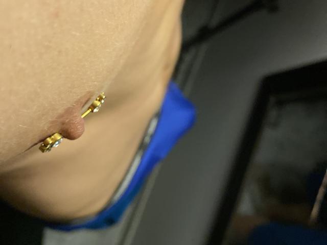 DeimHt's chest with a gold nipple piercing featuring small jewels, visible from the side. They're wearing blue underwear, showing a bit of their gray pants.