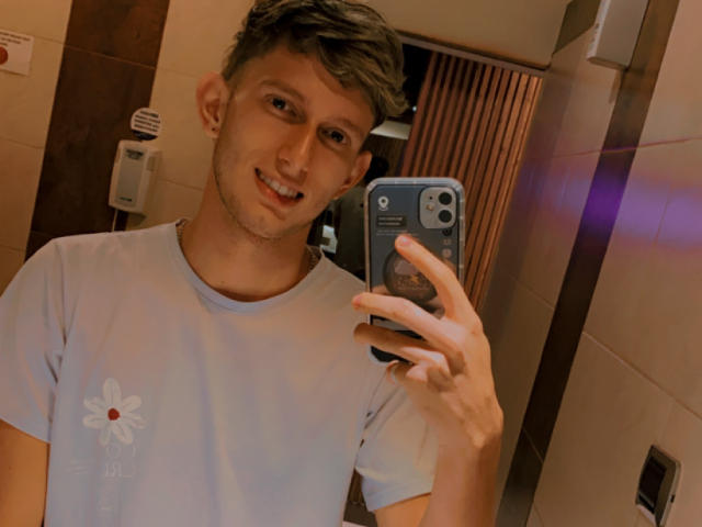 DeimHt with short brown hair, smiling, holding a phone in a mirror selfie, wearing a white T-shirt with a flower design, in a bathroom.