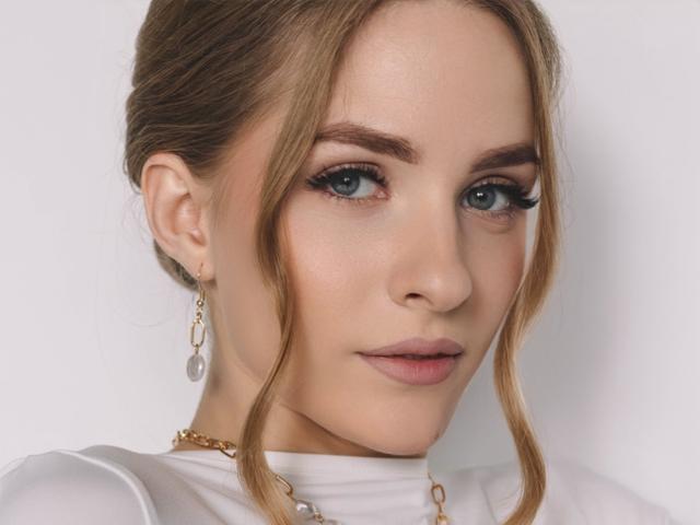 EvaDaly's face, showing her blue eyes, light makeup, and brown hair in a messy updo. She's wearing a white top, gold necklace, and silver earrings.