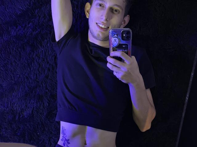 DeimHt lying on dark, fluffy carpet, taking a selfie with a blue-tinted phone, wearing a black crop top, showing a tattoo on their right side, smiling.