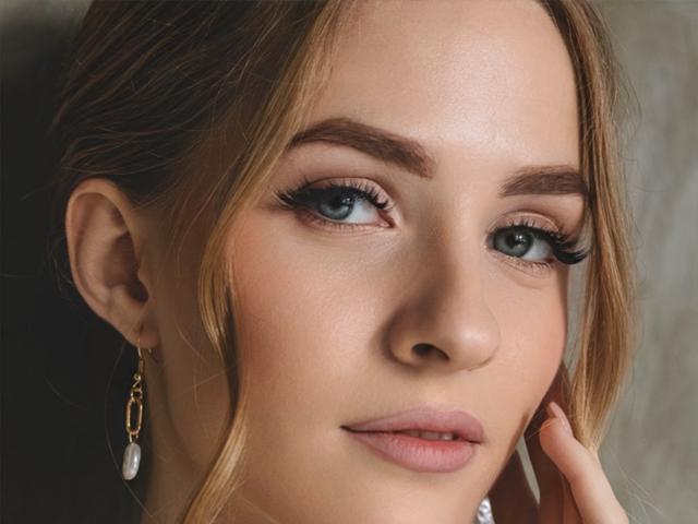 EvaDaly has blue eyes, light brown eyebrows, and long eyelashes. She's wearing gold earrings with white pearls, has pink lips, and a slight smile with her finger touching her cheek.