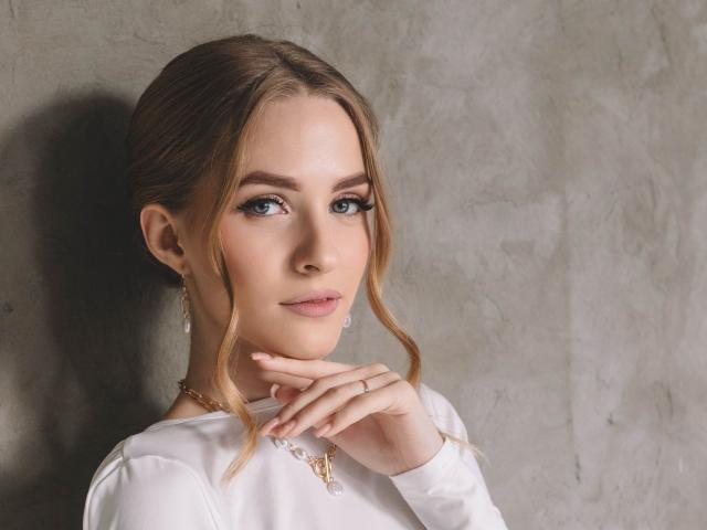 EvaDaly with blonde hair, blue eyes, and white shirt, touching her chin, wearing a gold necklace, and diamond earrings, looking at the camera with a subtle smile.