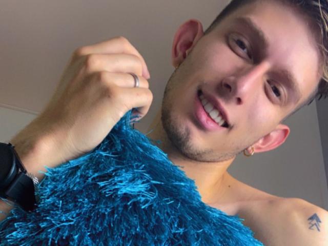 DeimHt holding a blue fluffy thing, shirtless, smiling with tongue out, short brown hair, small tattoo on left shoulder, gold earring, black watch, ring on right hand.