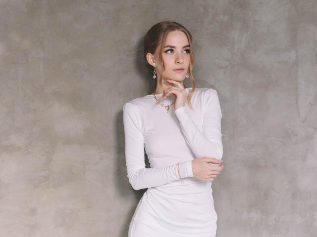 EvaDaly with blonde hair, white long-sleeve dress, holding chin and crossed arms, wearing earrings, soft smile, light skin, standing against textured gray wall.