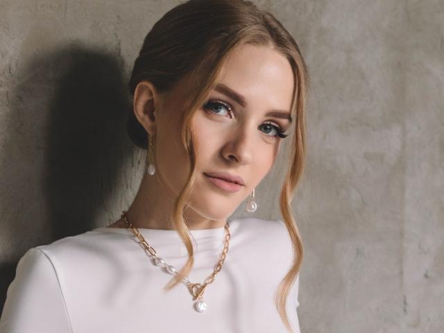 Eva Daly, con cabello rubio ondulado, ojos azules y una blusa blanca, lleva aretes de perlas y un collar de oro, está de pie frente a una pared de concreto gris, mirando ligeramente hacia un lado..
