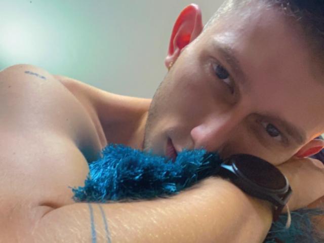 DeimHt, shirtless with a tattoo, holds a blue fuzzy thing to his mouth, wearing a black watch, red earbuds in, and staring intensely. Short brown hair, blue eyes.