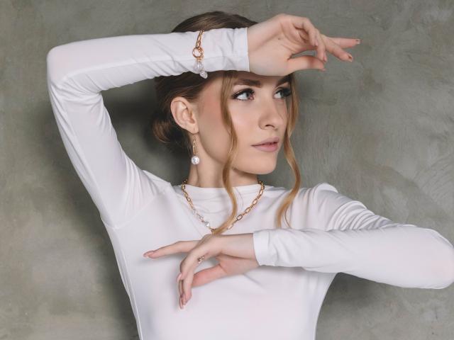 EvaDaly with blonde hair in a bun, wearing a white long-sleeve shirt, gold necklace, and pearl earrings, arms raised, looking to the side, showing her tits.