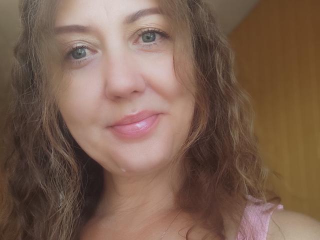 Close-up of RoseHille with wavy brown hair and light green eyes. She has a fair skin tone with a small droplet of saliva on her lower lip. She's wearing a pink sleeveless top. Her lips are slightly parted in a subtle smile. The image is well-lit with natural light highlighting her features. RoseHille's expression is relaxed and slightly playful. The background is a blurred yellowish area, keeping focus on her face.