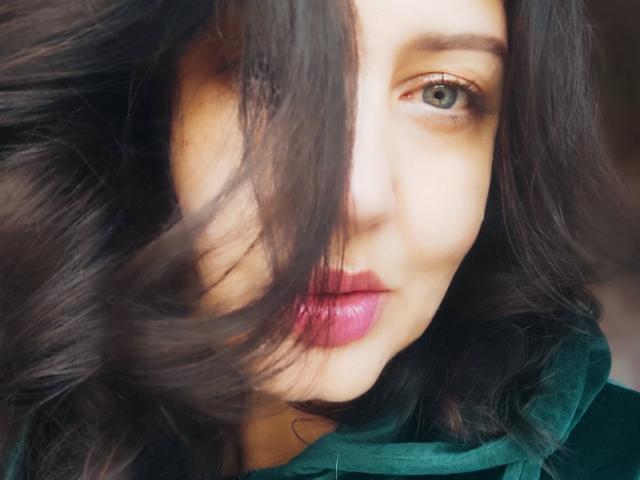 Close-up portrait of RoseHille with wavy dark brown hair partially covering her face. RoseHille has light skin, green eyes, and full pink lips. Her hair is slightly tousled, creating a dynamic effect. She wears a dark green garment with a soft texture. The light source is soft, highlighting her facial features. The image has a natural, slightly blurred style, emphasizing the movement of her hair.