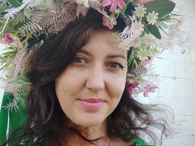 Close-up of RoseHille with wavy dark brown hair wearing a floral headpiece. The headpiece is full of pink and white flowers, with green leaves and delicate white lace. RoseHille has light brown eyes, pink lipstick, and a slight smile. She wears a green garment. The image focuses on her face, with the headpiece taking up most of the upper part of the frame. The texture of the flowers and lace contrasts with her smooth skin. The photo is brightly lit, highlighting her facial features and the intricate details of the headpiece.