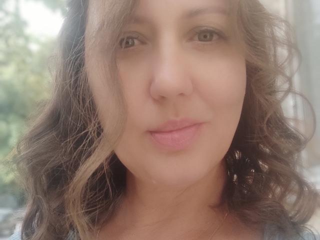 Close-up photo of RoseHille. She has light brown wavy hair, slightly parted to the right, with a soft, natural look. Her skin is smooth and fair, with subtle pink lips. RoseHille's brown eyes are centered, looking slightly to the left. She wears a thin gold necklace. The image is brightly lit with a slight blur in the background, emphasizing her face. Her expression is calm and neutral. The texture of her hair is soft and curly, contrasting with the smoothness of her skin.