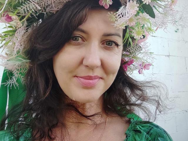 RoseHille with wavy dark brown hair, green eyes, pink lipstick, wearing a green dress and a flower crown with pink and white blossoms, smiling softly.