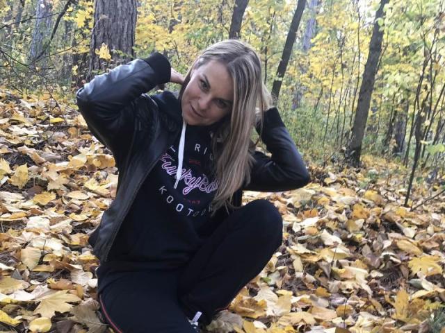 BelleLisaG with blonde hair, black hoodie, and black pants, squatting on yellow leaves, hands in hair, smiling, forest background, autumn.