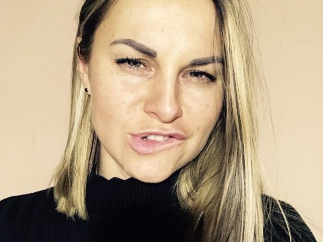 BelleLisaG has puffy pink lips, slightly squinted eyes, and blonde hair, wearing a black turtleneck, with a few small freckles on her face.