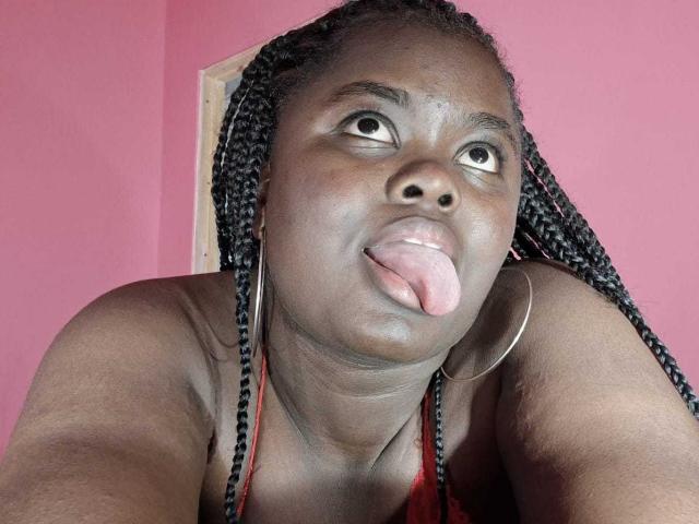 Close-up photo of ThickEbonyPrinces with dark skin and braided hair sticking out her tongue. She's wearing a red halter top and large hoop earrings. Her expression is playful, eyes looking slightly upwards. The pink background is simple and unobtrusive. The texture of her skin is smooth, and the braids are tightly coiled. The composition is centered, focusing on her face and upper torso. Her tongue is prominently out, adding a sense of humor and playfulness to the image.