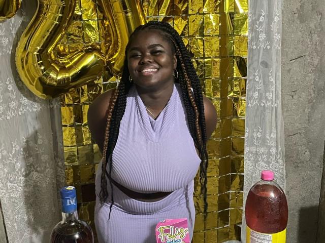 ThickEbonyPrinces with dark skin, long braided hair, big boobs, and a smile, wearing a purple halter top and matching pants, standing in front of gold balloons and a bottle.
