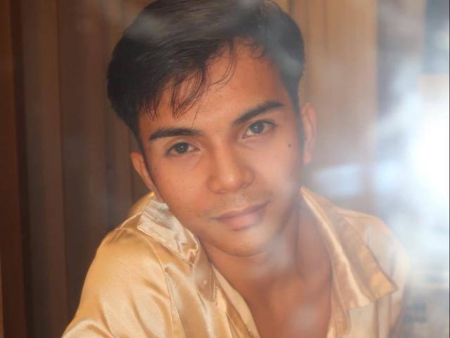 VanteLust has a cute, medium skin tone face with short black hair, wearing a shiny, beige shirt, looking softly at the camera with a slight, gentle smile.