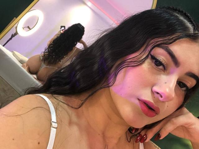 AlanaaWalker with wavy black hair, red lipstick, and white bra, looking at camera, while her butt and thong are reflected in a mirror behind her.