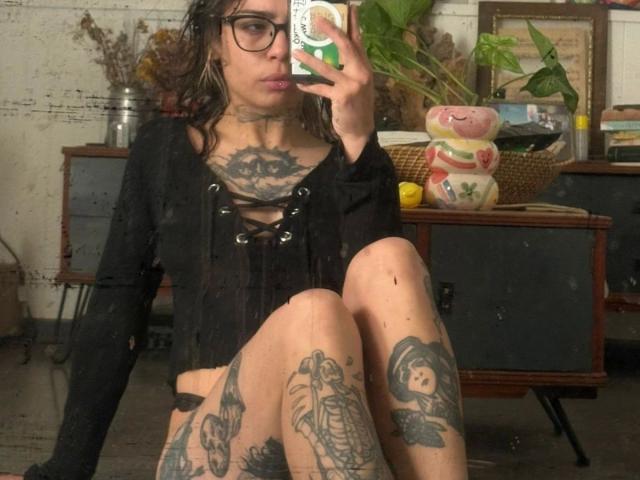 LunaRoses-hot with tattoos, wearing a black lace-up top, sitting with knees up, holding a phone, glasses, and wavy brown hair, showing her tattoos on chest and legs.