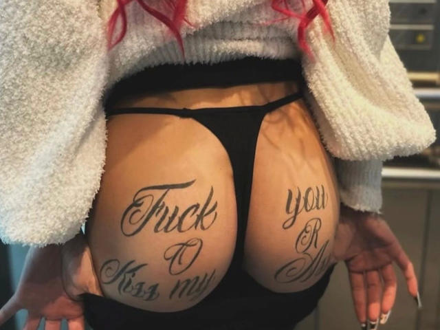MrsSkinnyGoddess-hot's ass, showing black thong, tattoos on both cheeks reading "Thick to Kiss me" and "You will" in cursive, with red hair and white fuzzy top.