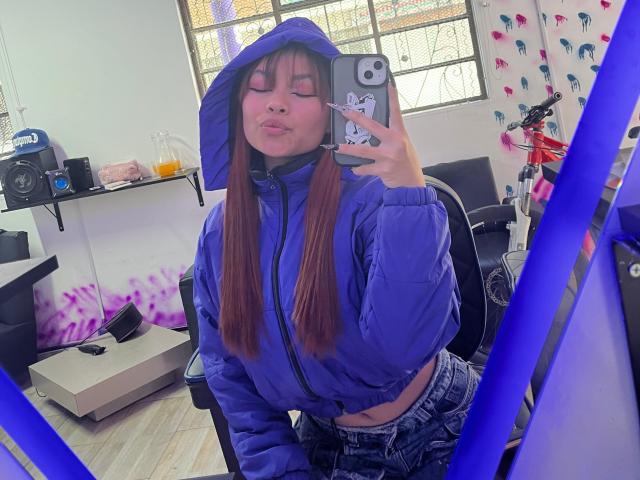 VannesaSaenz in a blue hoodie, pouting with her eyes closed, holding a phone with a cat sticker, showing her flat stomach and long brown hair. Mirror selfie.