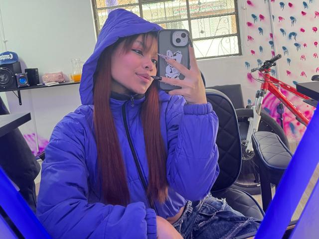 VannesaSaenz, a young woman with long brown hair, wears a blue puffy hooded jacket and denim jeans. She takes a mirror selfie with a phone featuring a Hello Kitty sticker. She's slightly sticking out her tongue. The room has a white wall, a window with glass blocks, a red bicycle, and colorful patterned decor. Her phone case is grey with a Hello Kitty sticker. The lighting is bright and even.