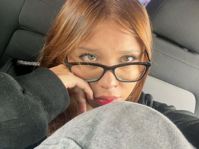 HeraJuno with brown eyes, black glasses, and red lipstick, resting her chin on a gray clothed thigh in a car, looking up with a slightly pouty expression.