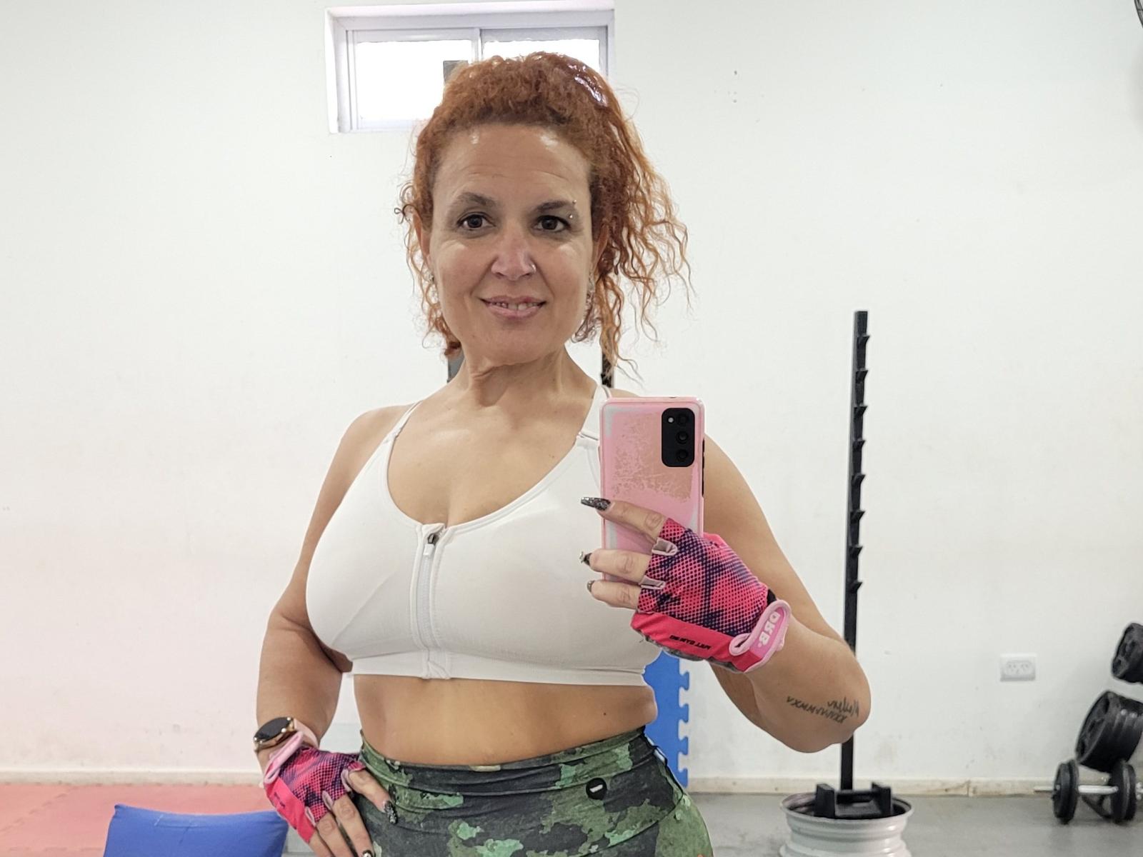 Colorulitos has curly red hair, wears a white sports bra, pink finger gloves, and green camo pants, holding a pink phone, smiling, showing her small tits and toned abs.