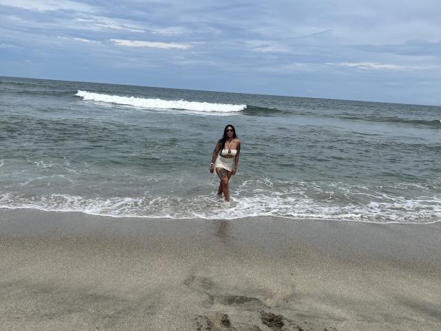 StellaBeckker stands in the shallow ocean, wearing a beige bikini, with small tits and a cute ass, long dark hair, and a slight smile, under a cloudy sky.