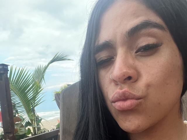 StellaBeckker has puffy lips, a nose ring, and long black hair, winking with slightly raised eyebrows. She's outside with a palm tree in the background.