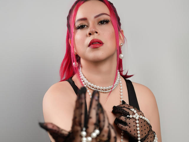 NatySilvy with pink hair, wearing pearl necklace, black lace gloves, and red lipstick, holding a pearl necklace in front of her, looking at the camera with a confident expression.