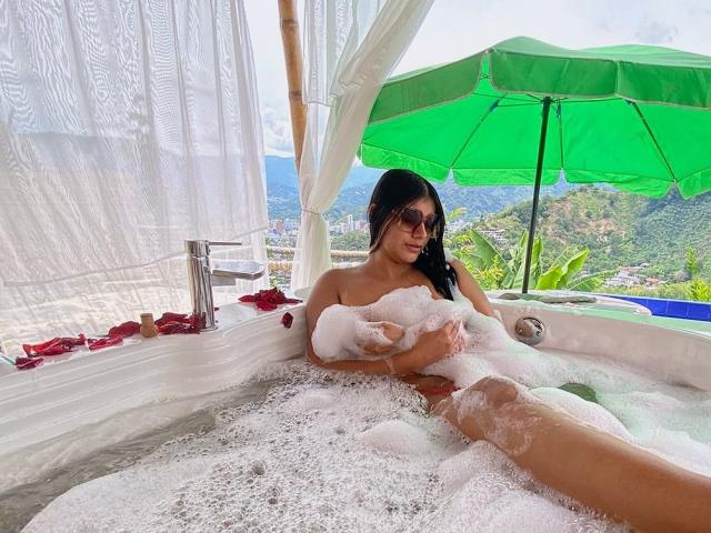 Abby Kell, con i capelli scuri, gli occhiali da sole e delle bolle di sapone che le coprono il seno, sdraiata in una vasca idromassaggio bianca con petali di rosa, un ombrello verde e una vista sulle montagne sullo sfondo..