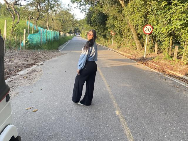 Abby Kell está em uma estrada, vestindo um suéter azul e preto com efeito degradê, calças pretas largas e sapatos verdes, com cabelos castanhos longos e um sorriso discreto..