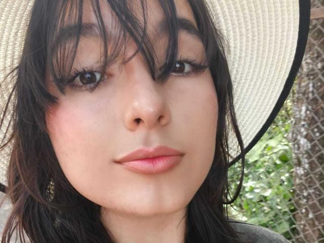 HairyValeri's face with dark, slightly wavy hair, wearing a wide-brimmed hat, has pink lips, subtle blush, and dark, half-closed eyes with long, thick eyelashes.