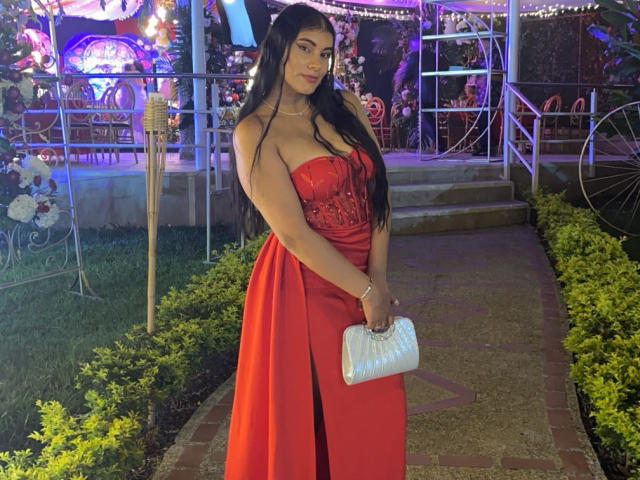 Abby Kell mit langem, schwarzem Haar, in einem roten, trägerlosen Kleid, das ihre Brüste zeigt, hält eine weiße Handtasche, steht auf einem Ziegelweg, mit Gras an den Seiten, im Abendlicht, und schaut in die Kamera..