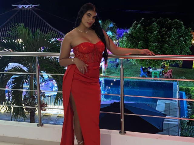 AbbyKell stands on a balcony, leaning on a metal railing. She's wearing a strapless, red, form-fitting gown with a high slit, revealing her white high-heeled sandals. Her long black hair flows over her right shoulder. She has a medium build with a curvy figure, large breasts, and a fair skin tone. Her right hand rests on the railing, and she looks directly at the camera with a slight smile. The background features a brightly lit pool area with greenery. The image has a night-time setting with artificial lighting.