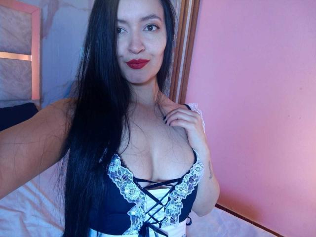 AriaBonnet with long black hair, red lipstick, and black lace-up lingerie, showing her tits, smiling, holding her top. Her hand is near her chest.