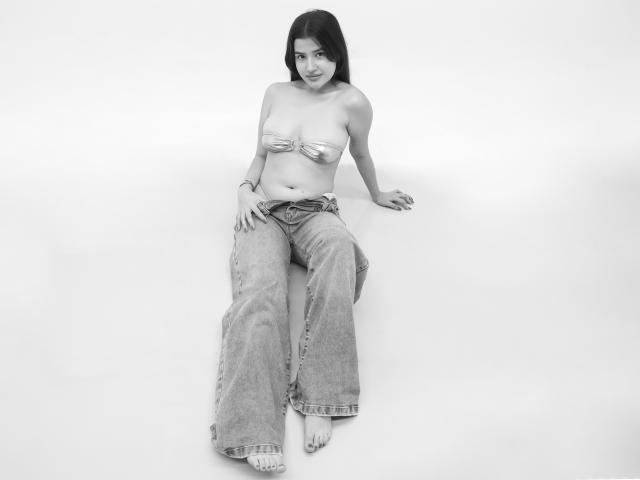 Iris Blair, con piccoli seni, indossa un top senza spalline lucido e jeans larghi, seduta a terra, sorridente, a piedi nudi e con i capelli scuri, in una foto in bianco e nero..