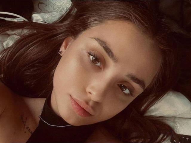 KerryCristal's face, with dark brown hair, green eyes, and a subtle smile, lying down, showing a small tattoo on her right shoulder. Soft, natural light.